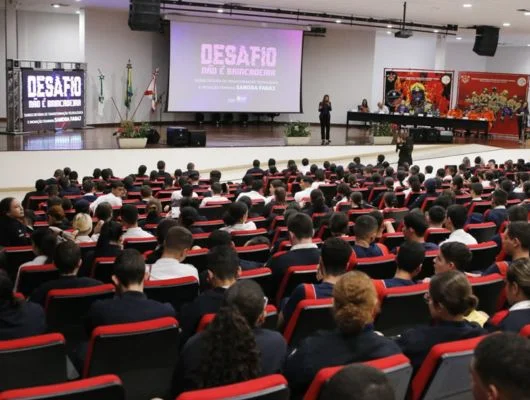 Sandra Faraj palestrando em auditório com a marca Desafio Não É Brincadeira projetada ao fundo